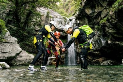 FP Events | Switzerland Tourism/Christian Meixner, un groupe de personnes en gilets de sauvetage et casques debout dans l’eau avec une cascade