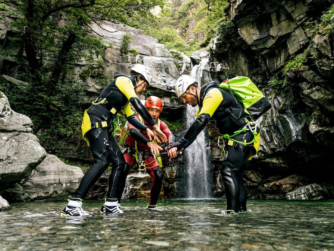 FP Events | Switzerland Tourism/Christian Meixner, un groupe de personnes en gilets de sauvetage et casques debout dans l’eau avec une cascade